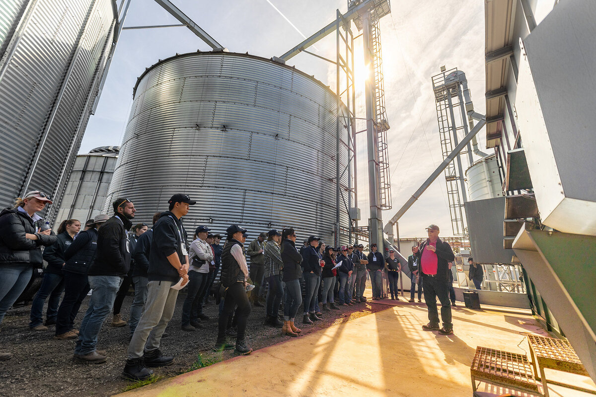 A large group of people gather around an Alvan Blanch grain dryer. They are surrounded by large feed silos